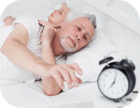 Ein älterer Mann liegt neben einer älteren Frau im Bett und streckt seine Hand Richtung Wecker